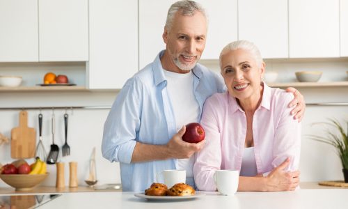 Image of cheerful mature loving couple family standing at the kitchen. Man holding apple. Looking at camera and hugging.