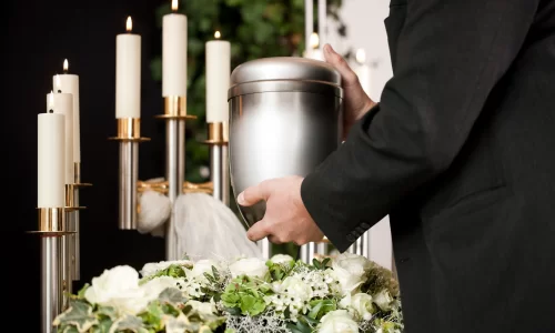Ambiente de funeralcom velas acesas, flores brancas e em primeiro plano o braço de uma pessoa vestindo paletó preto, segurando uma urna.
