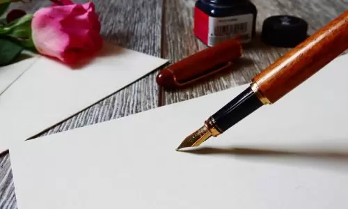 Sobre uma mesa, podemos ver uma caneta e papel, juntamente com um pote de tinta e uma flor